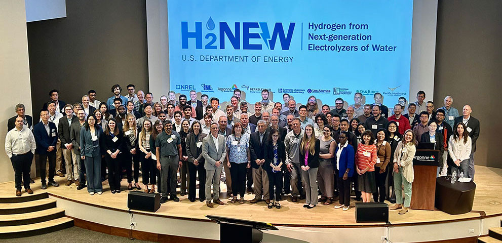 A large group of H2NEW researchers pose for a photo on an auditorium stage.