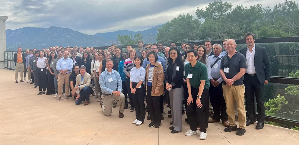 A large group of H2NEW researchers pose for a photo on an outdoor terrace with mountains in the background.